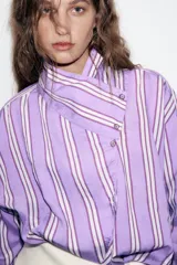 Camisa de corte oversize con diseño de rayas verticales en tonos violeta, blanco y bordó. Presenta cuello subido cruzado con cierre de botones frontal, mangas largas con puños y bajo asimétrico.