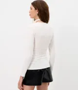 Blusa entallada de microfibra color blanco, con escote en V, mangas largas y detalle de frunces en los laterales. Presenta un lazo integrado en el cuello que permite anudarlo, aportando un toque sofisticado al diseño.