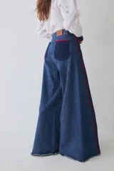 Jean de corte wide leg en color azul denim, con detalle de apliques de crochet en color rojo y azul a lo largo de las costuras laterales frontales. Presenta ruedo deshilachado y cierre frontal con botón metálico.