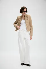 Blazer entallado de sarga color beige con cuello subido y escote en pico. Presenta manga larga con hombreras, detalle de botones metálicos decorativos en el frente y cierre frontal con gancho metálico. Incluye falsos bolsillos de vivo y abertura en la espalda.