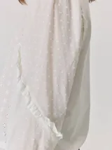 Camisa blanca de algodón con mangas largas abullonadas, detalle de volados en los hombros y puños elásticos. Presenta un diseño con cuello redondo y corte holgado.