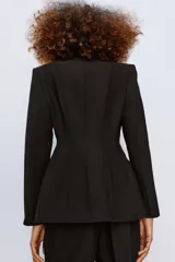 Blazer entallada de color negro con escote en pico, manga larga con hombreras y cierre frontal con botones ocultos. Presenta bolsillos con solapa en el delantero y detalle de nervios marcados en el tejido.