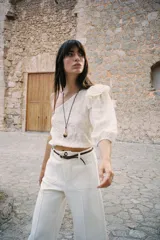 Top corto asimétrico color blanco roto, con un solo hombro descubierto y manga abullonada corta en el otro lado. Presenta bordados perforados (ojales) y un volante en el hombro descubierto. El bajo y el puño de la manga terminan en elástico.