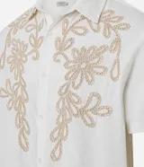 Camisa de corte comfort confeccionada en algodón, color blanco, con cuello solapa, manga corta y cierre frontal de botones. Presenta un delicado bordado floral en tono beige en la parte superior delantera.