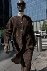 Abrigo marrón de gamuza con cuello redondo, manga larga, bolsillos delanteros y cierre frontal con botones.