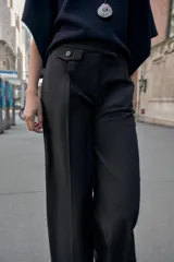 Pantalón de vestir marrón oscuro, tiro alto, con pernera ancha y pinzas frontales. Presenta un bolsillo con tapeta en la parte superior delantera.
