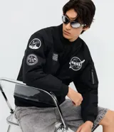 Campera bomber negra de microfibra, con cierre frontal por cremallera, puños y ruedo elásticos, y bolsillos con cierre. Cuenta con múltiples parches y estampas bordadas alusivas a la NASA, incluyendo el logo principal en el pecho, insignias en la manga derecha y una bandera de Estados Unidos.