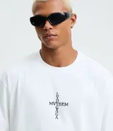 Remera blanca de algodón con modelado boxy, cuello redondo y manga corta. Presenta un estampado central en la parte delantera que forma una cruz con la palabra "MAYHEM" en el centro.
