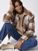 Sobrecamisa de estilo leñador con estampado de cuadros en tonos marrones, beige y gris. Presenta cuello clásico, cierre frontal con botones, bolsillos delanteros y puños tejidos acanalados. El interior está forrado con tejido tipo sherpa para mayor abrigo.