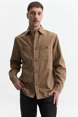 Sobrecamisa color marrón claro de twill, con cuello clásico, cierre frontal con botones a tono, mangas largas con puños abotonados, dos bolsillos tipo cartera con botón en el pecho y corte recto.