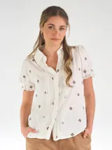 Camisa blanca de manga corta abullonada, con cuello camisero, cierre frontal con botones y detalles de encaje y bordados florales marrones.