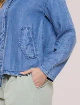 Camperita de denim liviano color azul, con cuello camisero, botones frontales ocultos, mangas largas de hombros caídos, bolsillos laterales y cordón ajustable en la cintura.