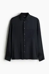 Camisa negra de satén con cuello inglés, tapeta lisa, bolsillo superior abierto y canesú en la espalda. Mangas largas con puños abotonados y bajo recto. Corte estándar.