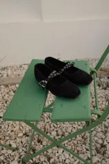 Ballerina de gamuza negra con puntera cuadrada y doble tira cruzada en el empeine con ojales metálicos y hebillas ajustables.