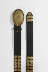 Cinturón de cuero vegano negro con hebilla grande de diseño irregular y acabado martillado en tono dorado. Presenta múltiples tachas circulares doradas dispuestas en dos filas a lo largo de la tira.