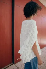 Top blanco de cuello redondo y mangas tipo capa, con textura fruncida, bajo asimétrico, lazos decorativos en el escote y en los laterales, y cierre posterior con botón.
