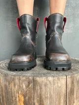 Bota de cuero con efecto desgastado en tonos marrones y negros, con suela track robusta y cierre lateral. Presenta detalles en color rojo en la parte superior de la caña.