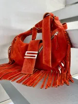 Cartera de cuero gamuza color naranja con diseño de media luna, decorada con flecos largos en los laterales. Presenta un cierre metálico superior, un herraje metálico distintivo en forma de S en el frente y un asa corta trenzada.