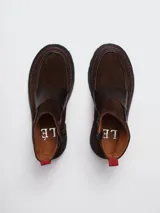 Botinetas de cuero y gamuza en tono marrón oscuro, con diseño de puntera reforzada y costuras decorativas en color rojo. Presentan una banda ancha sobre el empeine, cierre lateral y una lengüeta roja en el talón para facilitar el calce.