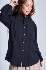 Camisa de color azul con cuello alto decorado con volados y lazo de ajuste. Presenta tablas decorativas en los hombros, cierre frontal con botones de nácar y mangas largas con puños elásticos que generan un efecto abullonado.