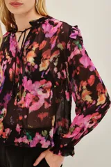 Camisa de viscosa con estampado floral en tonos fucsia, violeta y naranja sobre fondo negro. Presenta cuello con lazo ajustable y mangas largas con puños elásticos.