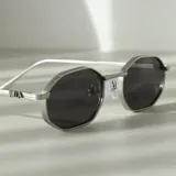 Lentes de sol con marco metálico plateado de forma octogonal y cristales oscuros. Presentan un diseño moderno con detalles estriados en el marco y el logo de la marca en las patillas.