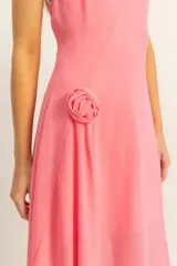 Vestido midi de color rosado con escote halter y espalda descubierta. Presenta un diseño asimétrico en el ruedo y un detalle de flor aplicada en la cintura.