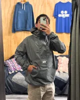 Rompeviento gris de estilo canguro con capucha, cuello alto y bolsillo frontal tipo sobre con cierre. Presenta puños elásticos y diseño funcional impermeable.