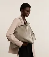 Bolso grande tipo hobo desestructurado, color gris claro con acabado ligeramente brillante o envejecido. Cuenta con dos asas cortas y una correa larga desmontable. Presenta detalles de cierres o tiradores decorativos en los laterales y cierre principal superior con cremallera.