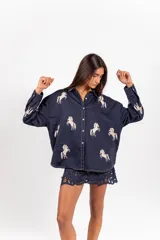 Camisa azul marino de corte oversize y manga larga, con cuello camisero, botonadura frontal y bordados contrastantes en crudo con motivos de caballos distribuidos en la prenda.