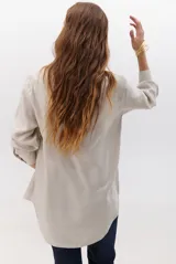 Camisa beige de manga larga con botones marrones y cuello clásico. Presenta un detalle de costura dorada a lo largo de la botonera.