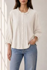 Blusa de manga larga color marrón, con cuello redondo y detalles fruncidos con volados pequeños en el cuello y los puños. Presenta cierre frontal con pequeños botones blancos y delicados pliegues verticales en el pecho.