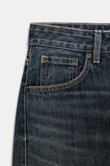 Jeans de tiro medio con diseño baggy balloon, cinco bolsillos y costuras laterales torcidas. Presenta un acabado desgastado con roturas localizadas y cierre frontal con cremallera y botón metálico.