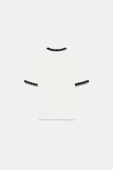 Camiseta blanca de manga corta y cuello redondo, con ribete negro decorado con perlas blancas en el cuello y los puños.
