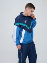 Campera deportiva Umbro modelo Beat, confeccionada en nylon ligero con diseño de bloques de color en blanco, azul marino y verde azulado. Presenta cierre frontal, cuello alto y elásticos en puños y cintura.