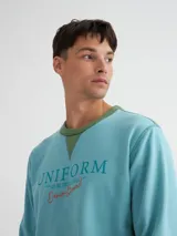 Buzo azul con cuello redondo, mangas largas y puños y bajo acanalados. Presenta bloque de color naranja en hombros y laterales, y estampado frontal con texto en color borgoña.