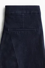 Jean de tiro alto con corte acampanado desde la rodilla. Presenta un diseño de denim oscuro con pliegues frontales y traseros, cierre con botón oculto y bolsillos traseros decorativos.