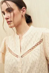 Camisa de algodón color beige con cuello mao, cierre frontal de botones y mangas cortas con volados. Presenta un diseño texturizado con rayas verticales y detalles de pasamanería calada en el frente.