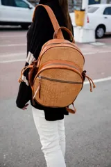 Mochila de cuero color tostado con textura de reptil en los bolsillos frontales. Cuenta con compartimento principal con cierre, bolsillos frontales con cierre, tiras ajustables y cordones laterales decorativos.