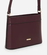 Cartera bandolera pequeña de formato rectangular, confeccionada en poliuretano con acabado liso. Presenta una correa larga ajustable, cierre superior con cremallera y un pequeño aplique metálico dorado con el logo de la marca en el frente.