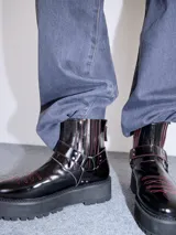 Bota corta de cuero negro tipo biker, con suela track de goma, plataforma, costuras y bordados decorativos en hilo rojo. Presenta tiras con hebillas metálicas y cierre en la parte trasera.