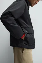 Parka de corte relajado en color negro, confeccionada en tejido de algodón con interior ligeramente acolchado. Presenta cuello de solapa en pana, cierre frontal con cremallera y bolsillos delanteros tipo plastrón con solapa y botones a presión.