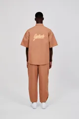 Sobrecamisa unisex color marrón claro, de manga corta y corte ancho. Presenta abertura frontal con botones tono sobre tono, dos bolsillos de parche en la parte inferior delantera y un bolsillo de ribete en el pecho. Logotipo de la marca estampado en contraste en el pecho y estampado grande en dos tonos en la espalda.