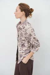 Camisa de manga larga con cuello clásico y cierre frontal de botones. Presenta un estampado floral en tonos marrones sobre una base color beige, confeccionada en un tejido con textura similar al gamuzado.