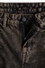 Jean de denim de algodón rígido, color negro con efecto lavado ácido o desgastado en tonos marrones/óxido. Modelo de tiro medio/estándar, con cinco bolsillos y costuras frontales marcadas que recorren la pierna. Presenta una abertura frontal en el bajo de cada pierna.