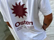 Remera blanca de corte oversize con cuello redondo y manga corta. Presenta un estampado frontal en color marrón rojizo con un diseño abstracto que incluye un sol estilizado y la palabra "Osten".