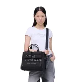 Bolso tipo tote Marc Jacobs modelo The Cargo Canvas Medium, color negro. Confeccionado en lona resistente, cuenta con asas superiores, correa ajustable para el hombro y múltiples bolsillos delanteros externos. Presenta el texto "THE TOTE BAG" y el logo de la marca bordados en el frente.