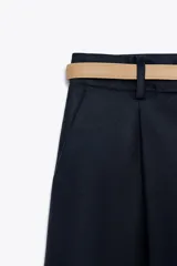 Bermuda de tiro medio color azul marino, con pinzas delanteras, cierre frontal y cinturón fino color beige.