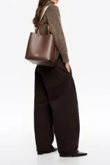 Bolso de mano tipo tote de Bimba y Lola, color marrón, confeccionado en piel reconstituida. Presenta doble asa de mano, asa de tejido ajustable con logo de la marca, cierre de botón de presión y logo metálico frontal.