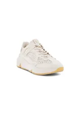 Zapatilla deportiva estilo chunky, color beige claro, con paneles superpuestos de cuero y perforaciones decorativas. Presenta una suela gruesa y voluminosa de color blanco con detalles en goma natural en la punta y el talón.